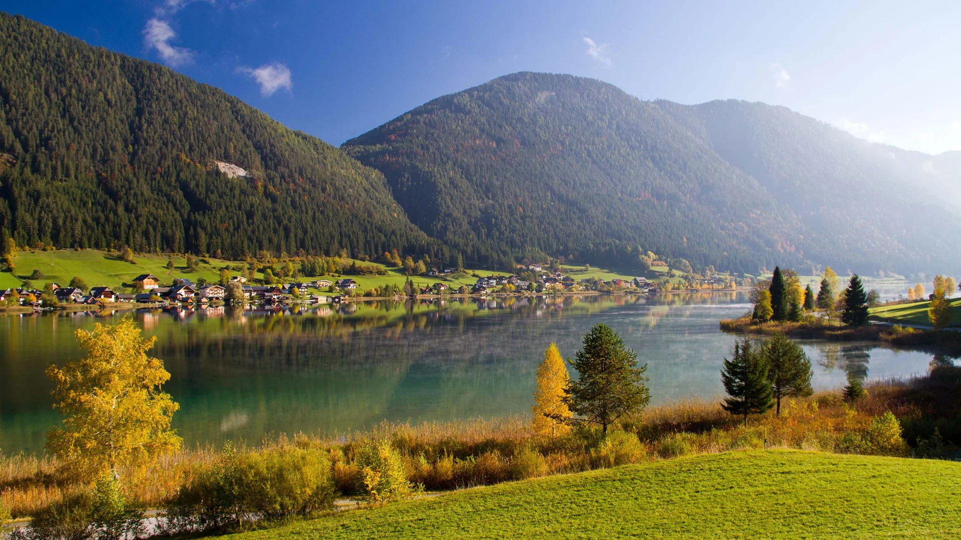 Bergsee mit Dorf, bewaldeten Hängen und herbstlicher Landschaft