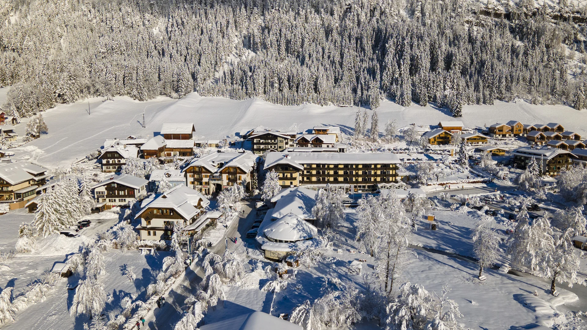 Verschneites Dorf mit Häusern vor bewaldeten Bergen im Winter