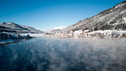 Winterlandschaft mit verschneitem Dorf an einem nebligen See und Bergen