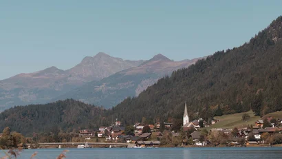 See mit Dorf und Kirchturm vor bewaldeten Bergen und Schilf im Vordergrund