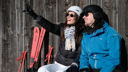 Paar in Skibekleidung lehnt an Holzwand, Frau zeigt auf etwas