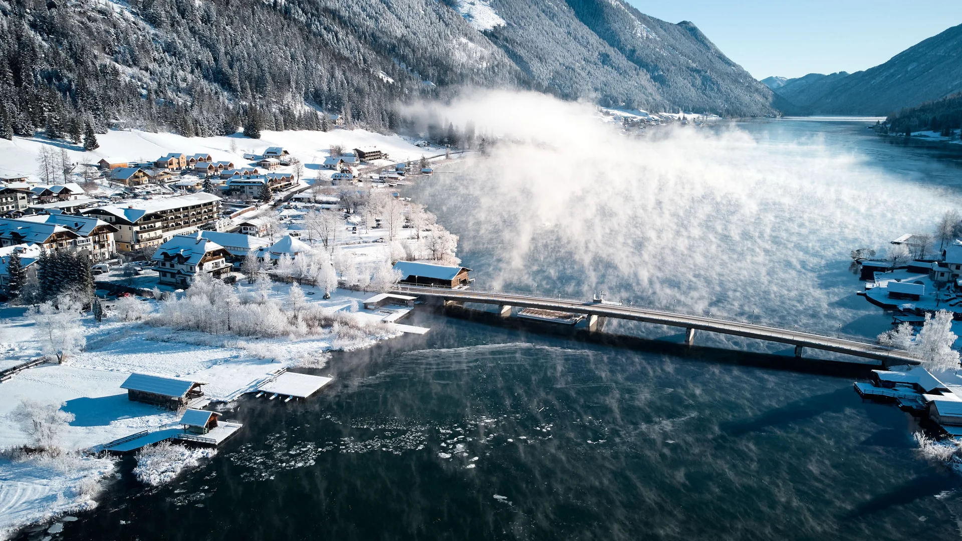 Winterlandschaft mit verschneiten Häusern, Steg und dampfendem See in den Bergen