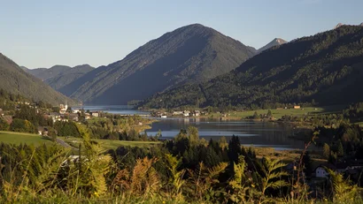 Blick auf einen Bergsee, ein Dorf und bewaldete Berge