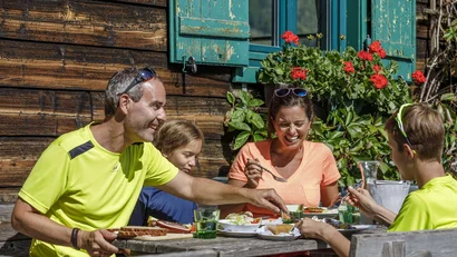 Familie isst gemeinsam an einem Holztisch vor einem rustikalen Holzhaus