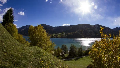 Bergsee mit bewaldeten Bergen und Herbstlaub unter blauem Himmel