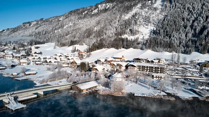 Verschneites Dorf am See vor bewaldetem Berg unter blauem Himmel