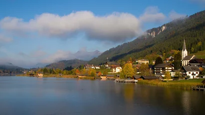 Seeufer mit Dorf, Kirche und bewaldeten Bergen im Hintergrund