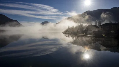 Nebeliger Bergsee mit Uferhäusern und Spiegelung im Morgenlicht