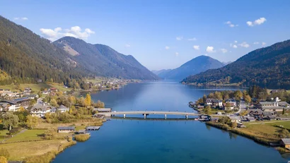 Bergsee mit Brücke, Dörfern und bewaldeten Bergen unter blauem Himmel