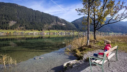Person auf Bank am See mit Bergpanorama und Dorf am Ufer